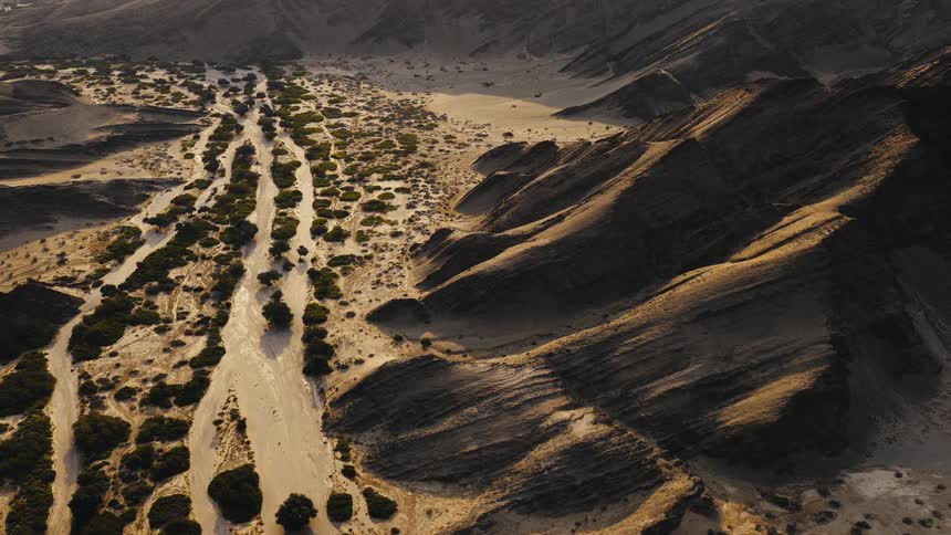 A vast desert valley where green shrubs grow along a dry riverbed between steep rocky mountains during sunset