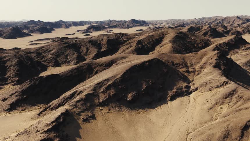 An aerial sweep reveals a vast rugged desert landscape dominated by dark rocky ridges and golden sand dunesthe camera glides over the arid terrain showcasing the sharp contrast between the jagged mountain peaks and the smooth desert floor below