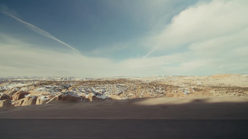 A view from a moving vehicle shows a vast desert landscape with rocky cliffs and light snow under a bright blue sky with thin clouds