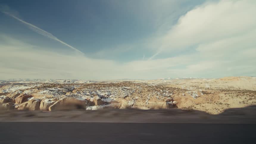 A view from a moving vehicle shows a vast desert landscape with rocky cliffs and light snow under a bright blue sky with thin clouds