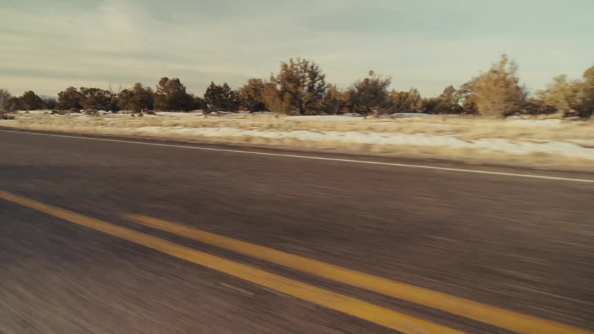 A roadside view shows a road with a solid yellow line and a field with scattered trees and snow