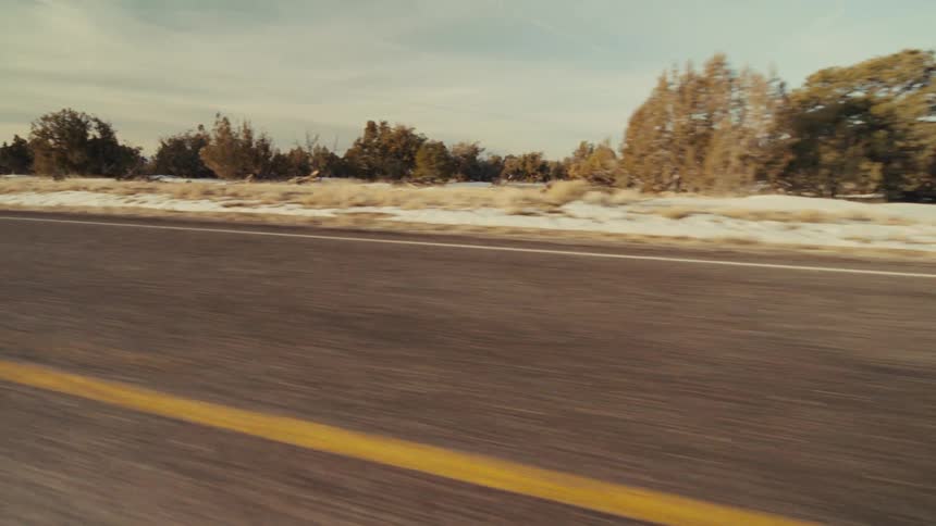 A roadside view shows a road with a solid yellow line and a field with scattered trees and snow