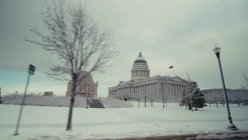 The camera pans past a neoclassical capitol building with a prominent dome set against a grey overcast skythe surrounding grounds and a nearby stone monument are blanketed in a thick layer of fresh snow