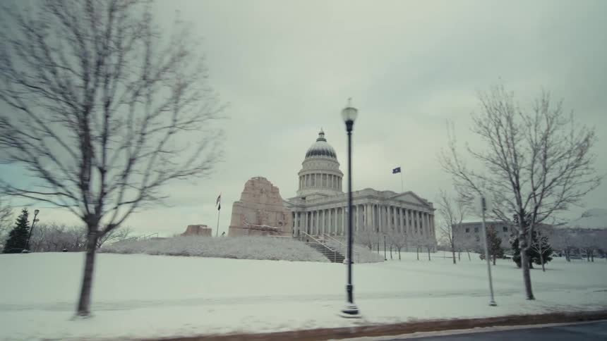 The camera pans past a neoclassical capitol building with a prominent dome set against a grey overcast skythe surrounding grounds and a nearby stone monument are blanketed in a thick layer of fresh snow