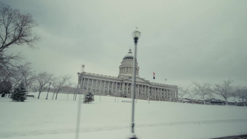 A view from a moving vehicle shows a large stone capitol building with a dome situated on a snowy hill behind bare trees under a grey sky
