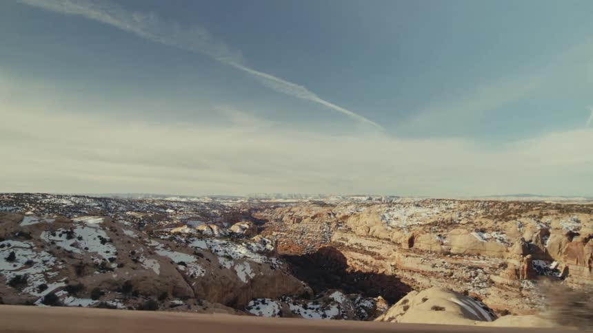 A view from a moving vehicle shows a vast desert landscape with rocky cliffs and patches of white snow under a blue sky with thin clouds