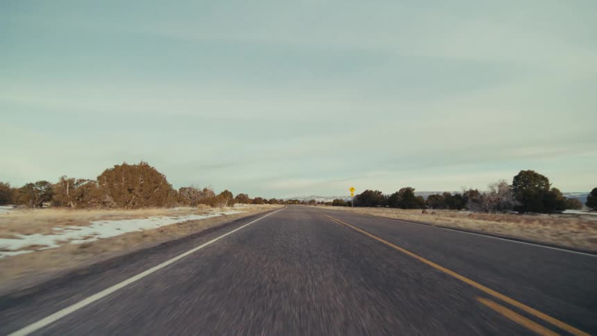 A long asphalt road through a desert landscape under a pale sky with low shrubs and yellow traffic signs lining the shoulder while mountains appear in the distance