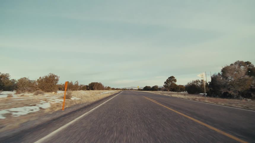 A long asphalt road through a desert landscape under a pale sky with low shrubs and yellow traffic signs lining the shoulder while mountains appear in the distance