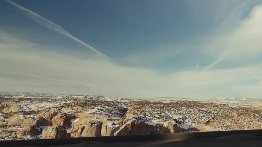 A wide shot of a desert landscape with scattered snow featuring rock formations and a vast sky with contrails