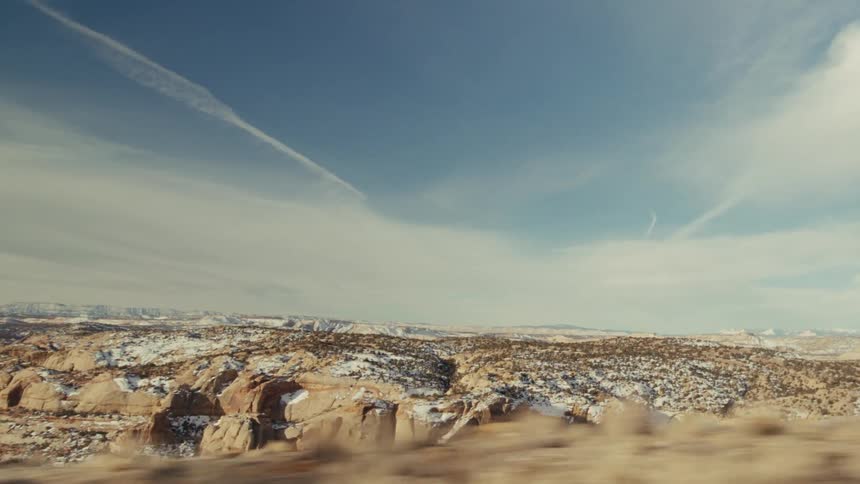 A wide shot of a desert landscape with scattered snow featuring rock formations and a vast sky with contrails