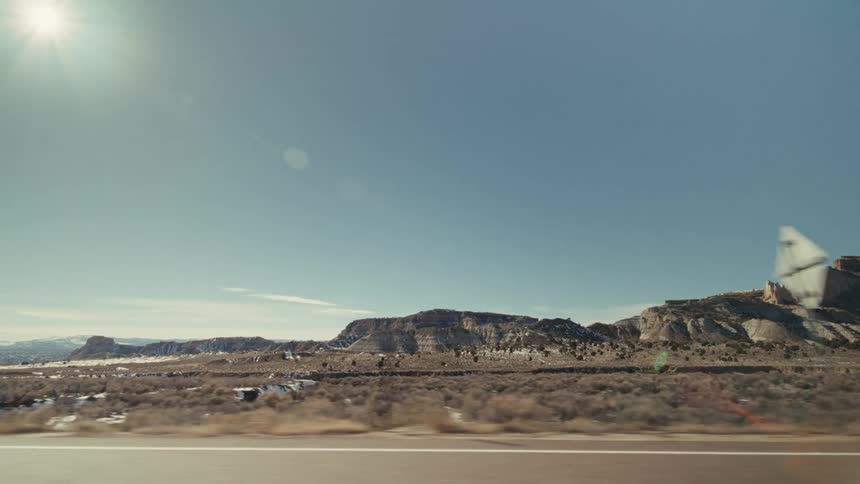 A vast desert landscape from a moving car while passing rocky mesas and sparse vegetation under a bright sun and clear blue sky