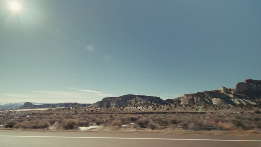 A vast desert landscape from a moving car while passing rocky mesas and sparse vegetation under a bright sun and clear blue sky