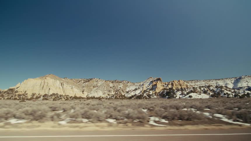 The camera travels alongside a vast desert landscape featuring lightcolored rocky mesas and patches of snow under a clear blue sky lowlying shrubs cover the foreground as the road stretches past the distant rugged mountain formations