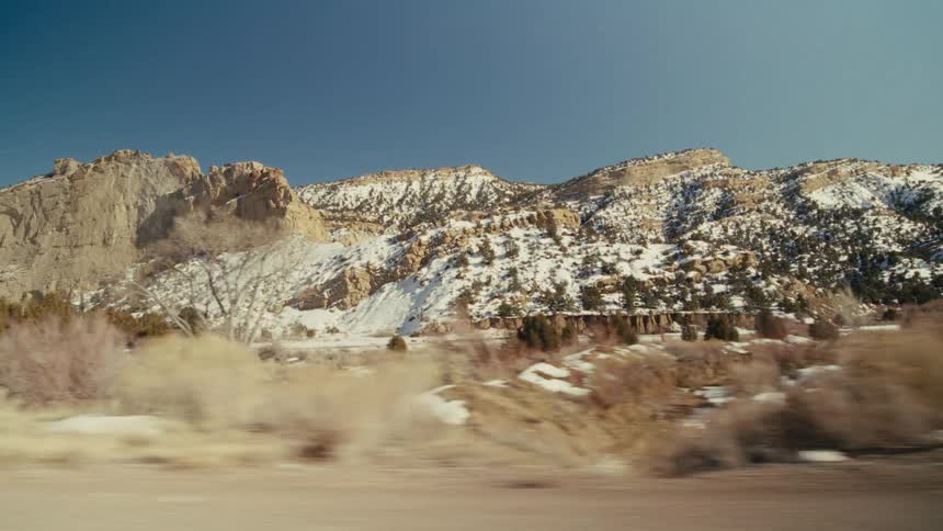 A vast desert landscape from a moving vehicle showing large rocky mountains covered in patches of snow under a clear blue sky with dry brush and scattered trees in the foreground