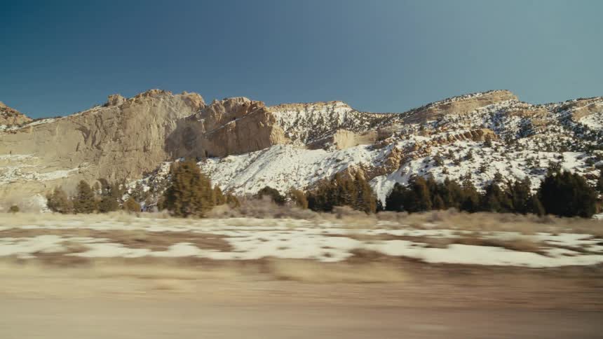 A vast desert landscape from a moving vehicle showing large rocky mountains covered in patches of snow under a clear blue sky with dry brush and scattered trees in the foreground