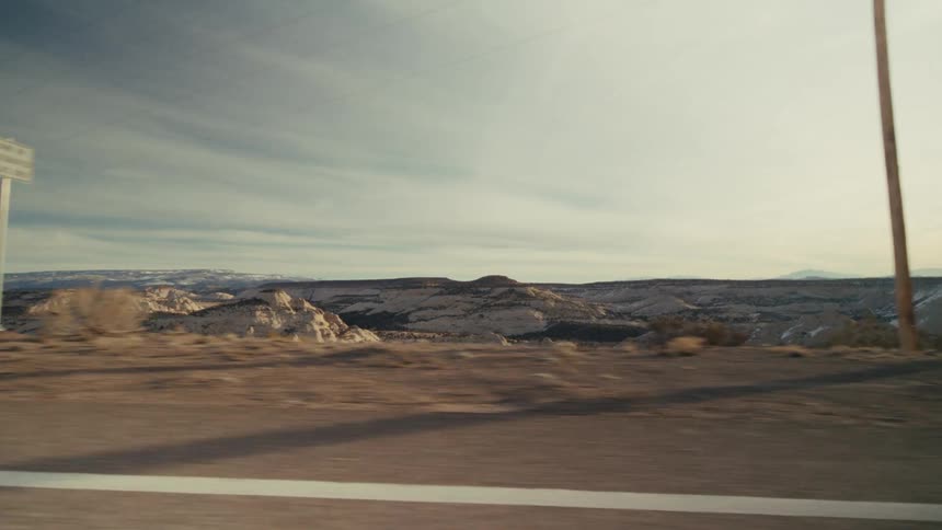 Videoa view from a moving vehicle shows a wide desert valley with beige rocky hills and scattered green bushes under a soft blue sky with light clouds