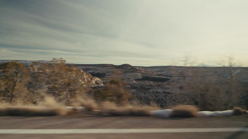 Videoa view from a moving vehicle shows a wide desert valley with beige rocky hills and scattered green bushes under a soft blue sky with light clouds