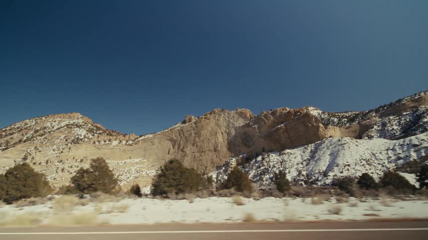 A view from a moving vehicle travels along a straight paved road through a dry landscape with light brown rocky mountains and scattered green trees under a clear blue sky