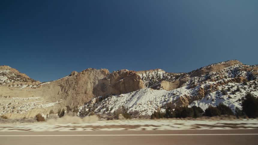 A view from a moving vehicle travels along a straight paved road through a dry landscape with light brown rocky mountains and scattered green trees under a clear blue sky