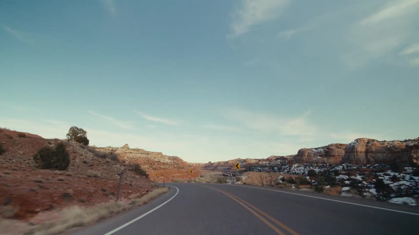 A car travels along a winding twolane highway that cuts through a vast desert canyon landscapethe surrounding terrain features red rock formations and sparse vegetation with patches of snow visible on the distant hillsides under a clear blue sky