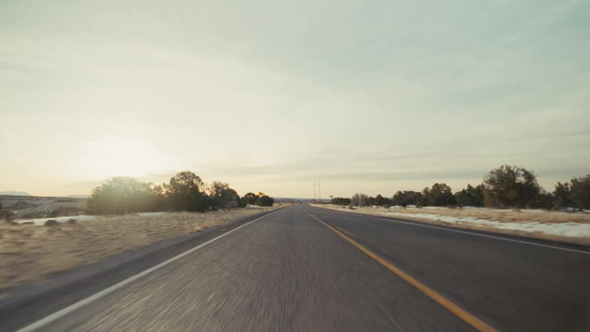 A view from a moving vehicle travels down a long straight road with two yellow lines through a dry landscape with scattered trees and patches of snow under a soft evening sky