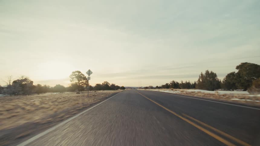A view from a moving vehicle travels down a long straight road with two yellow lines through a dry landscape with scattered trees and patches of snow under a soft evening sky