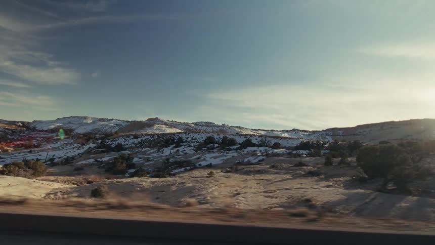 A view from a moving vehicle shows a vast desert landscape with patches of white snow on rocky hills under a bright blue sky with thin clouds