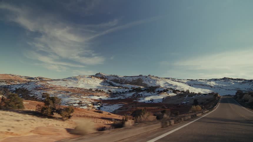 A view from a moving vehicle shows a paved road winding through a vast desert valley with snow covered hills and rocky mountains under a blue sky with soft white clouds