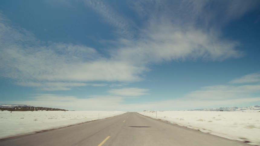 A long twolane highway stretches toward the horizon through a vast snowcovered landscape under a bright blue sky with wispy cloudsthe camera provides a forwardmoving perspective along the road where low hills and distant mountains are