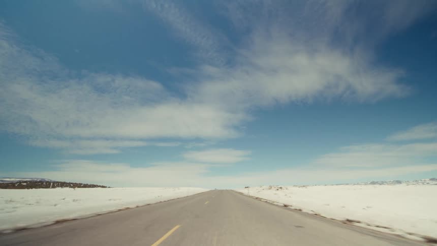 A long twolane highway stretches toward the horizon through a vast snowcovered landscape under a bright blue sky with wispy cloudsthe camera provides a forwardmoving perspective along the road where low hills and distant mountains are
