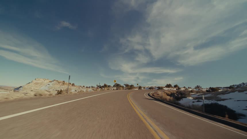 A view from a moving vehicle travels along a curved paved road with two yellow lines through a rocky landscape with patches of white snow under a bright blue sky with light clouds