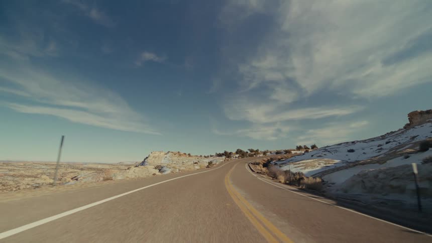 A view from a moving vehicle travels along a curved paved road with two yellow lines through a rocky landscape with patches of white snow under a bright blue sky with light clouds
