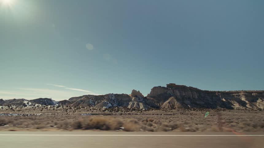 The video shows a sideview perspective from a moving vehicle traveling along a highway through a vast desert landscape large rugged rock formations and mesas sit under a clear blue sky with the bright sun creating a slight lens flare in the upper left