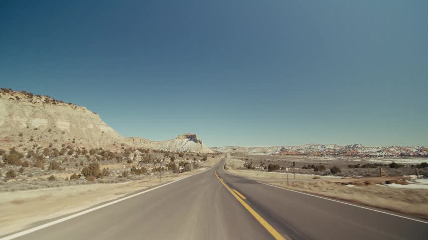 A view from a moving vehicle travels along a straight paved highway with yellow lines through a dry desert valley featuring white rocky hills and small bushes under a clear blue sky