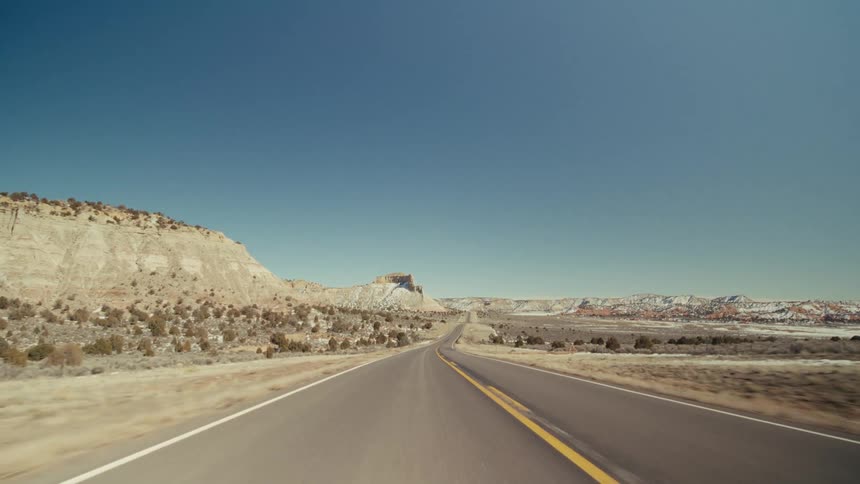 A view from a moving vehicle travels along a straight paved highway with yellow lines through a dry desert valley featuring white rocky hills and small bushes under a clear blue sky