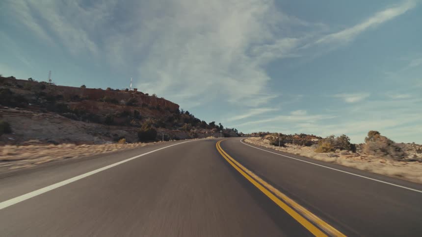 A view from a moving vehicle travels along a curved paved highway with two yellow lines through a rocky canyon with patches of white snow under a blue sky with soft clouds