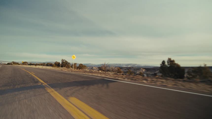 A paved two lane highway stretching through a dry desert landscape under a pale sky with distant hills and sparse vegetation visible along the roadside