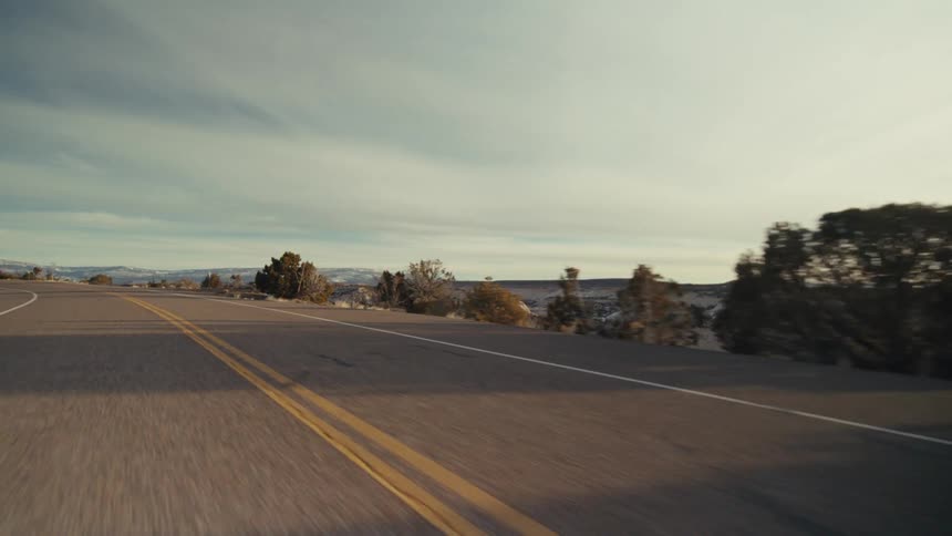 A paved two lane highway stretching through a dry desert landscape under a pale sky with distant hills and sparse vegetation visible along the roadside