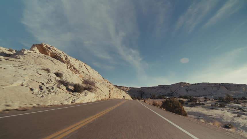 A curved two lane highway through a vast desert canyon under a bright sun with scattered snow covering the rocky hillsides and sparse vegetation under a blue sky
