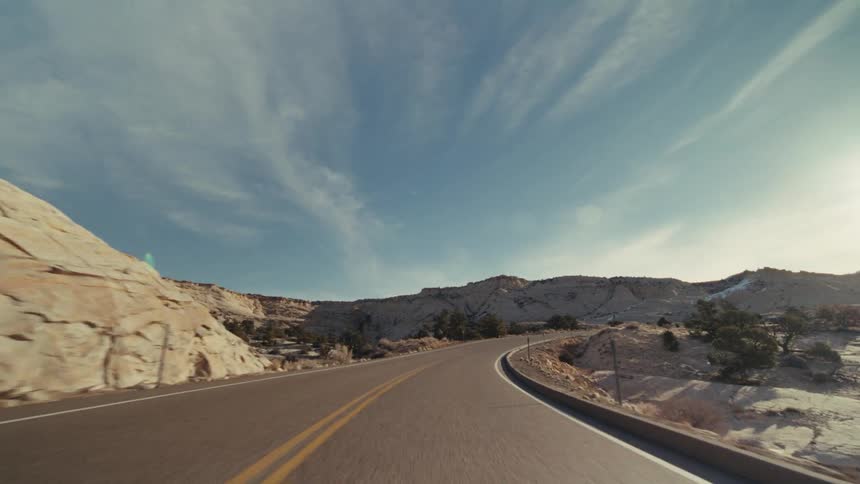 A curved two lane highway through a vast desert canyon under a bright sun with scattered snow covering the rocky hillsides and sparse vegetation under a blue sky