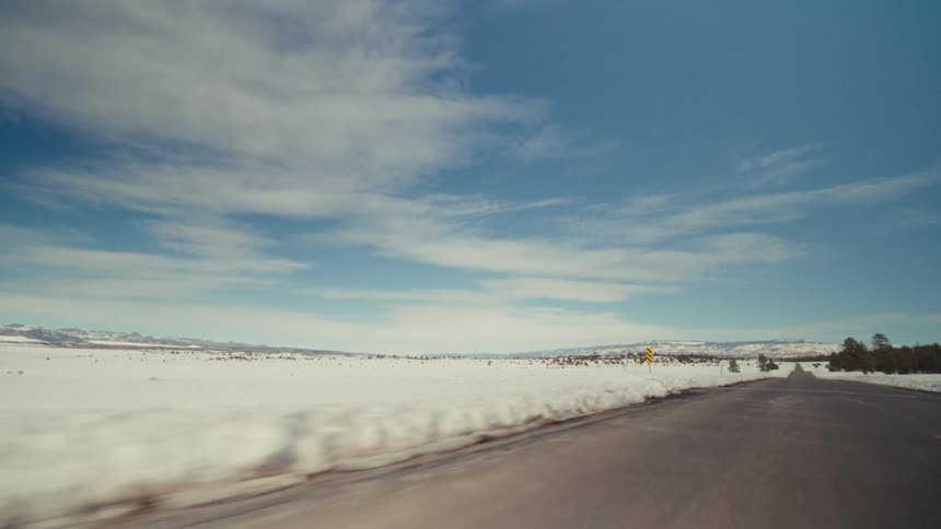 A view from a moving vehicle shows a long paved road passing through a vast snowy field with scattered evergreen trees under a bright blue sky with soft white clouds