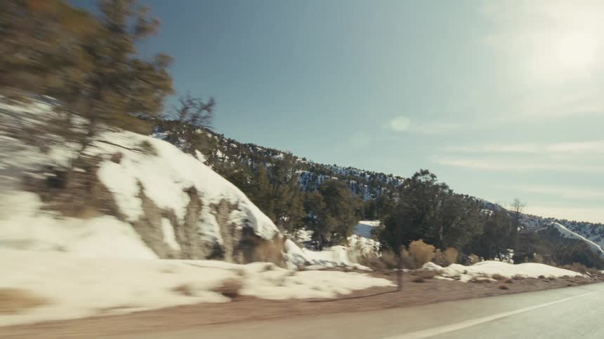 A view from a moving vehicle travels along a curved paved highway with two yellow lines past steep snowy hills and evergreen trees under a bright sun and clear blue sky
