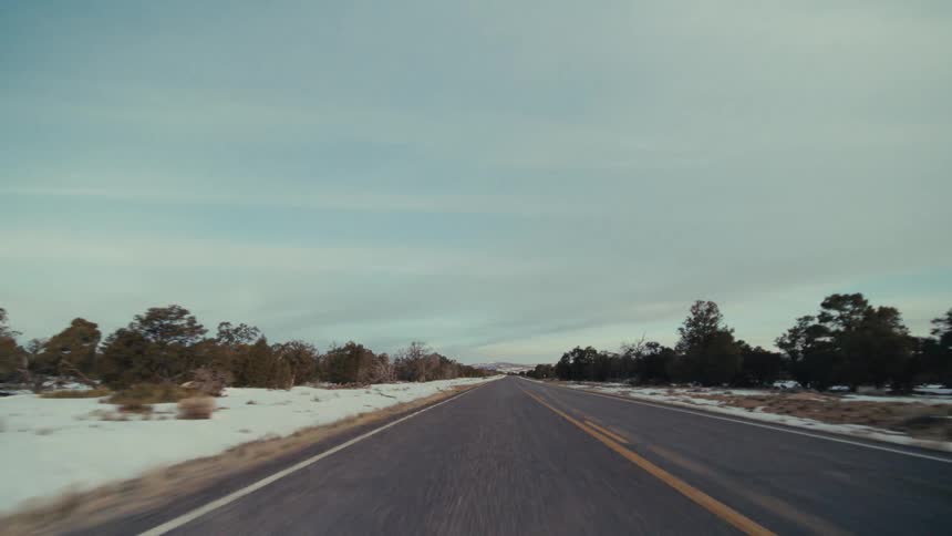 A point of view shot from a vehicle driving on a two lane road through a snowy tree lined landscape under a cloudy sky