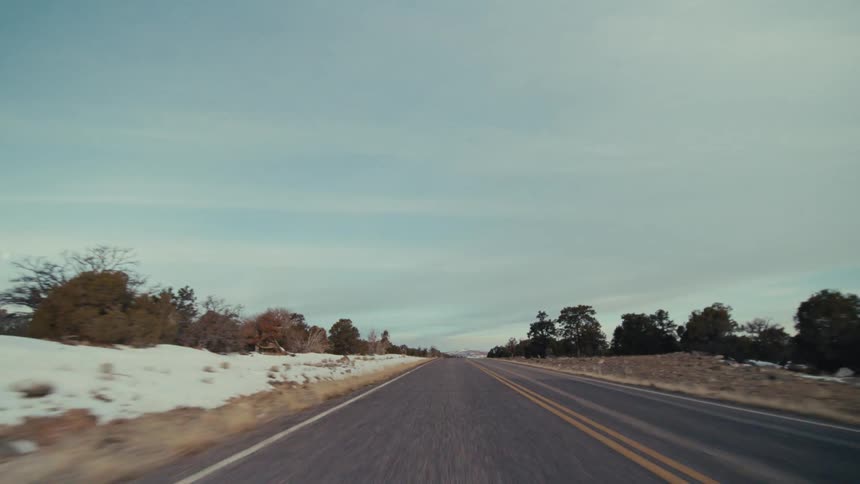 A point of view shot from a vehicle driving on a two lane road through a snowy tree lined landscape under a cloudy sky