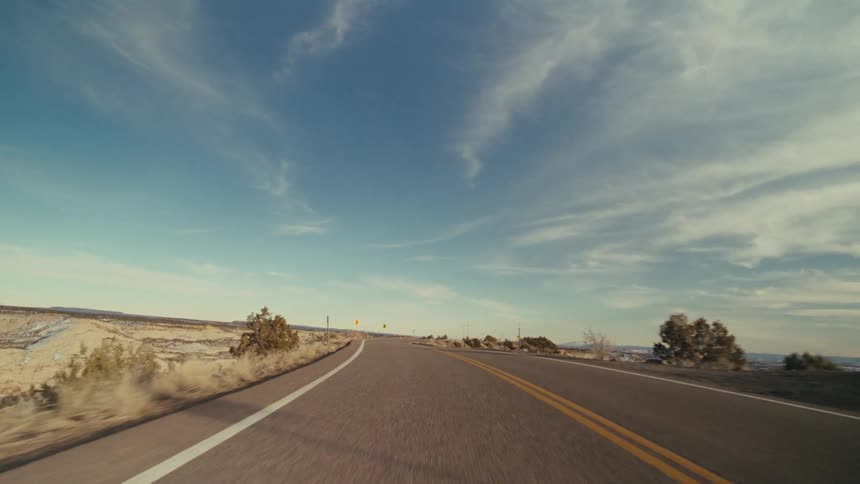 A view from a moving vehicle shows a paved road winding through a vast desert valley with snow covered hills and rocky mountains under a blue sky with soft white clouds