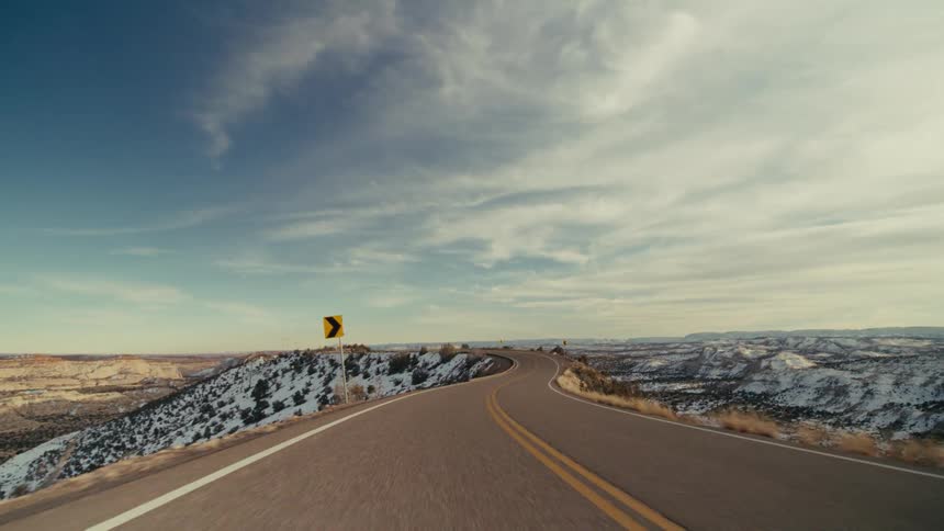 A view from a moving vehicle shows a paved road winding through a vast desert valley with snow covered hills and rocky mountains under a blue sky with soft white clouds