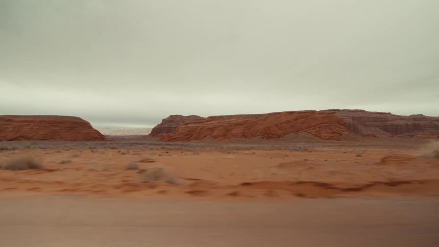 A view from a moving vehicle shows a paved road passing through a vast orange desert landscape with large rock formations under a grey overcast sky