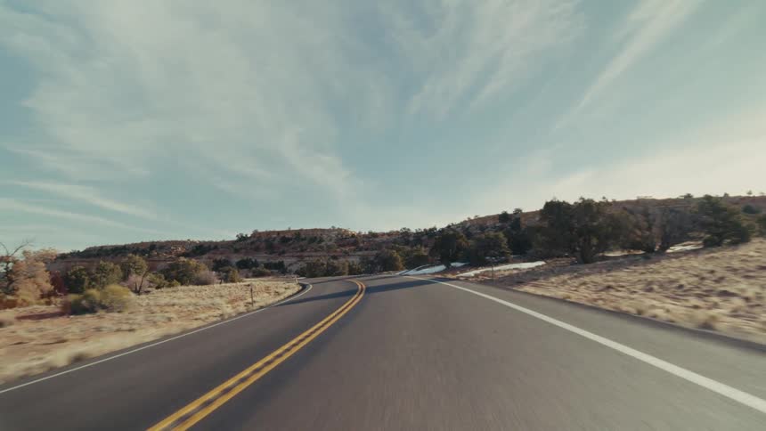 The video captures a forwardfacing pointofview from a vehicle driving along a winding desert highway the bright sun shines over a vast arid landscape featuring rocky hillsides dotted with patches of snow and sparse vegetation
