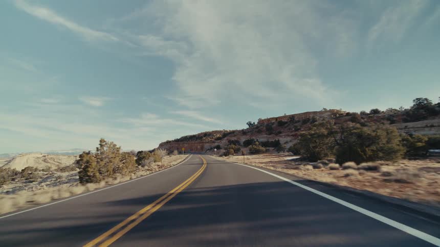 The video captures a forwardfacing pointofview from a vehicle driving along a winding desert highway the bright sun shines over a vast arid landscape featuring rocky hillsides dotted with patches of snow and sparse vegetation
