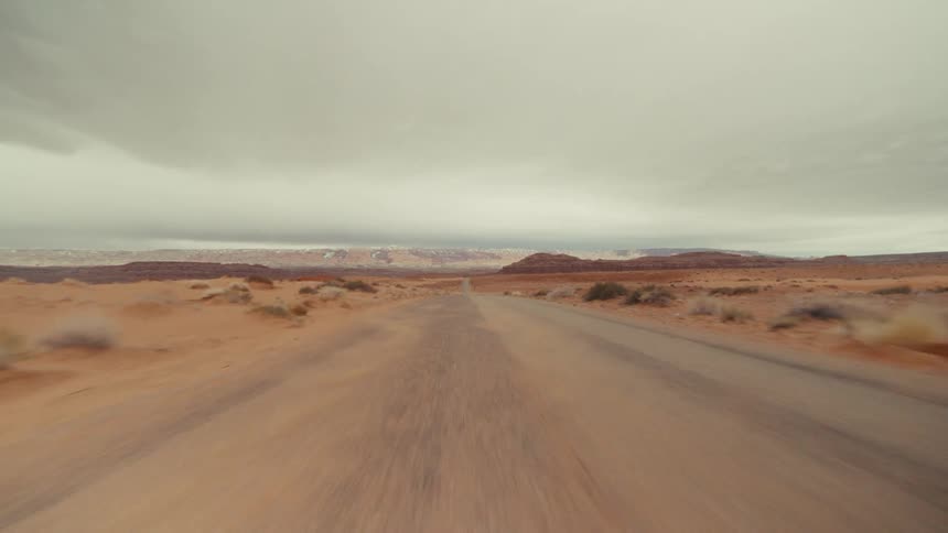 A point of view shot shows a vehicle driving down a straight desert road under an overcast sky